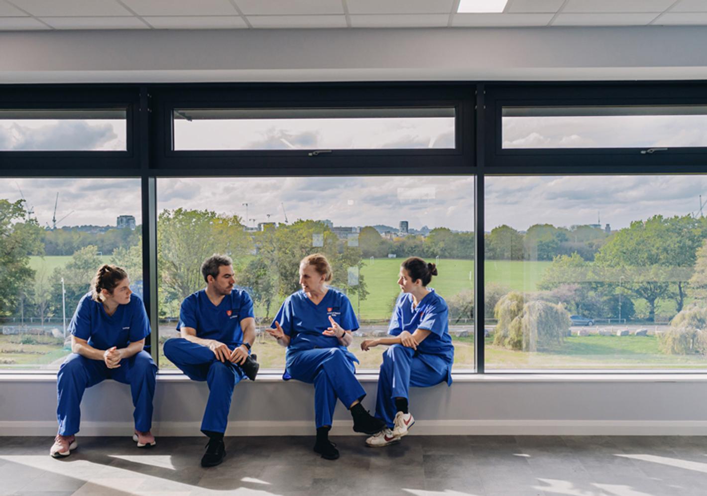A group of medical professionals in scrubs sit talking in front of a large window that looks out on to green countryside