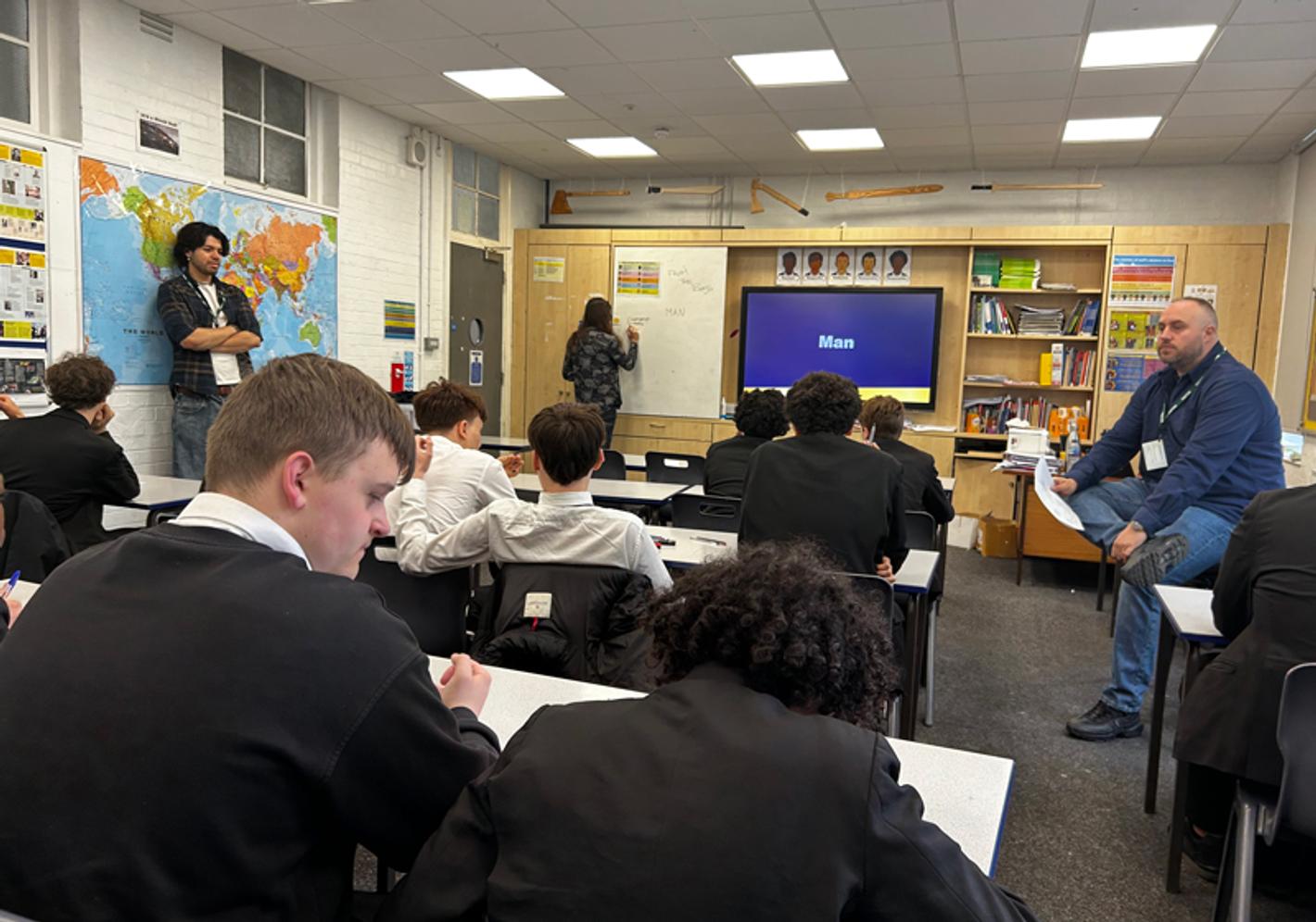 A group of young males on a classroom watching a teacher write on a whiteboard