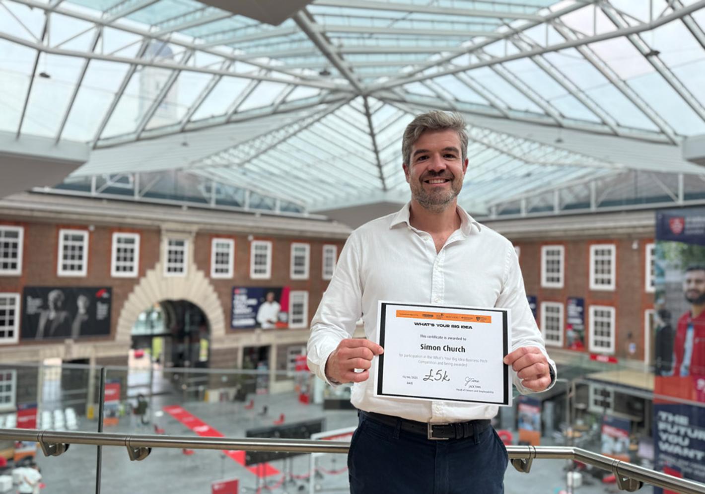 A 91��ɫ MBA student in a white shirt stands holding a certificate on the second floor balcony of the Quad