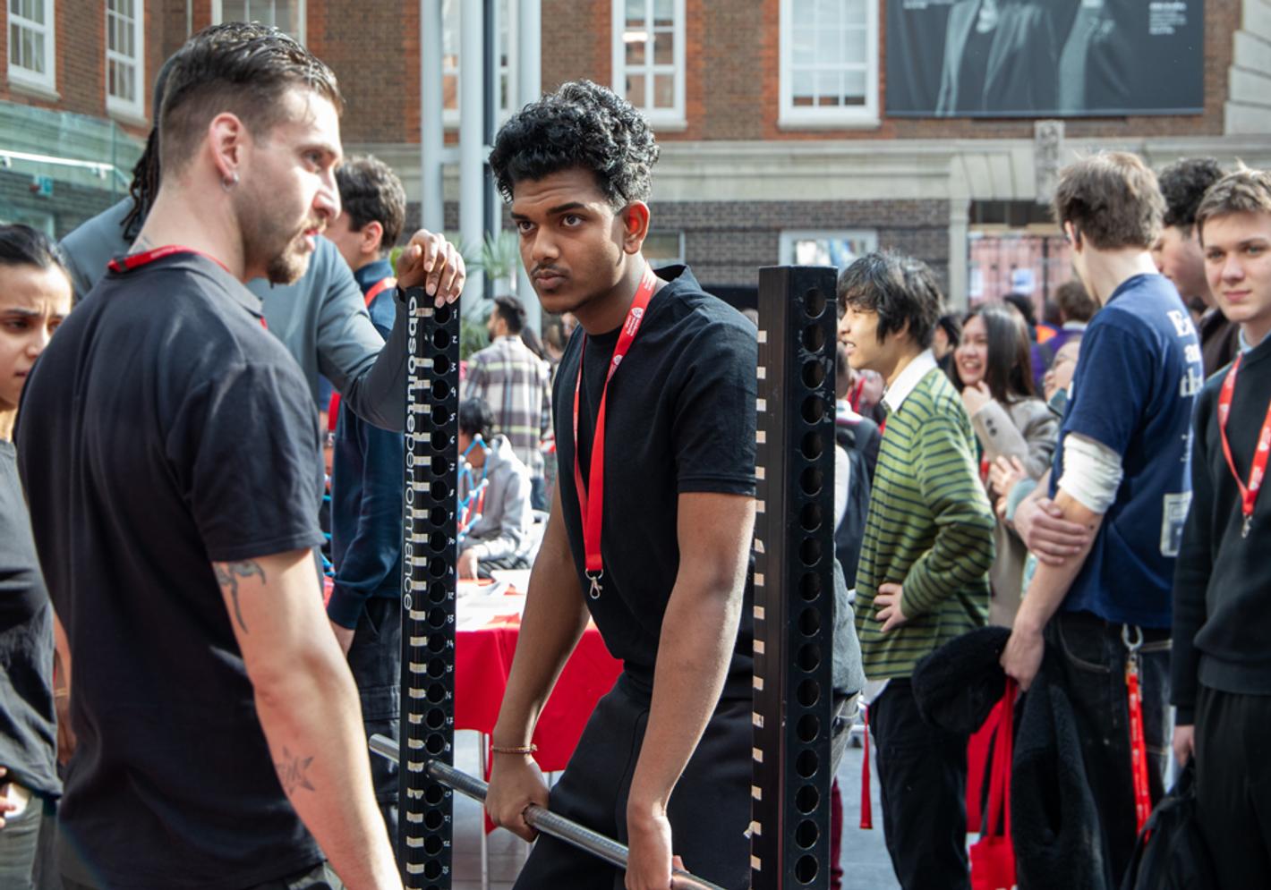 A young man lifts weights under supervision at a crowded event