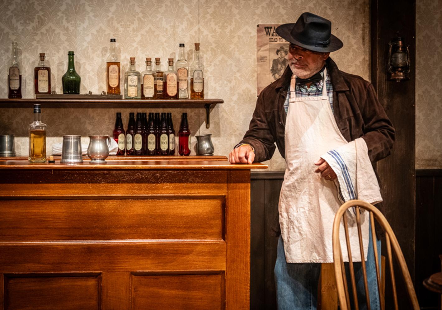 A man wearing a hat and apron leans against a bar with drinks in the background