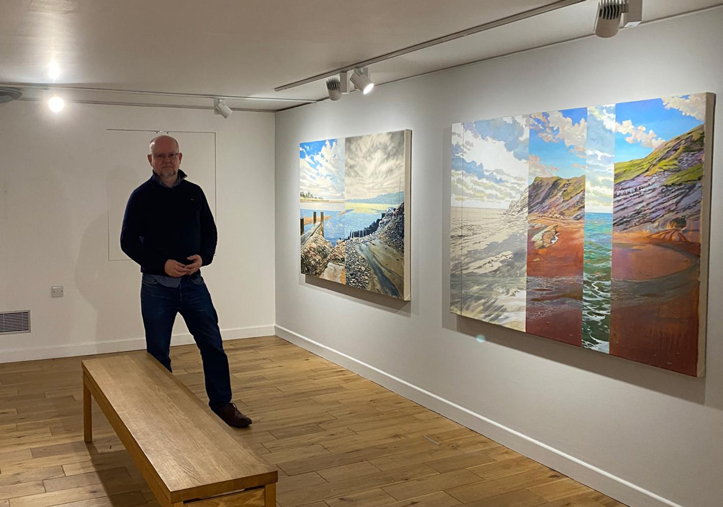 A portrait of Dr John Timberlake standing next to his paintings in the Granary Gallery, Berwick-Upon-Tweed