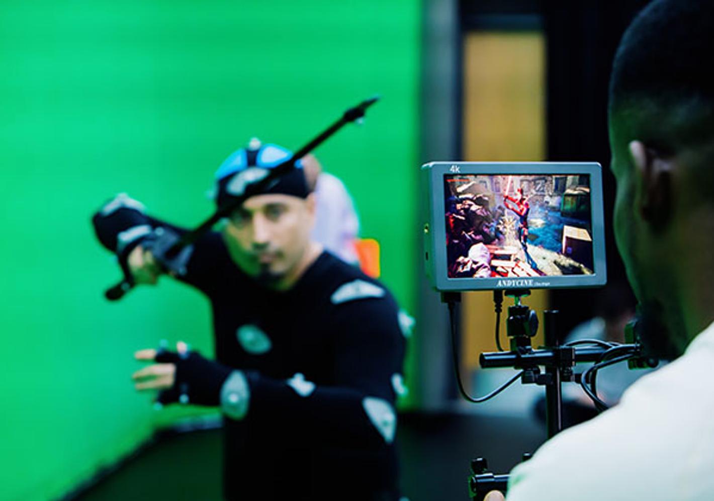 student holds sword in front of green screen while another looks into camera viewfinder where first student is seen in CGI landscape and costume