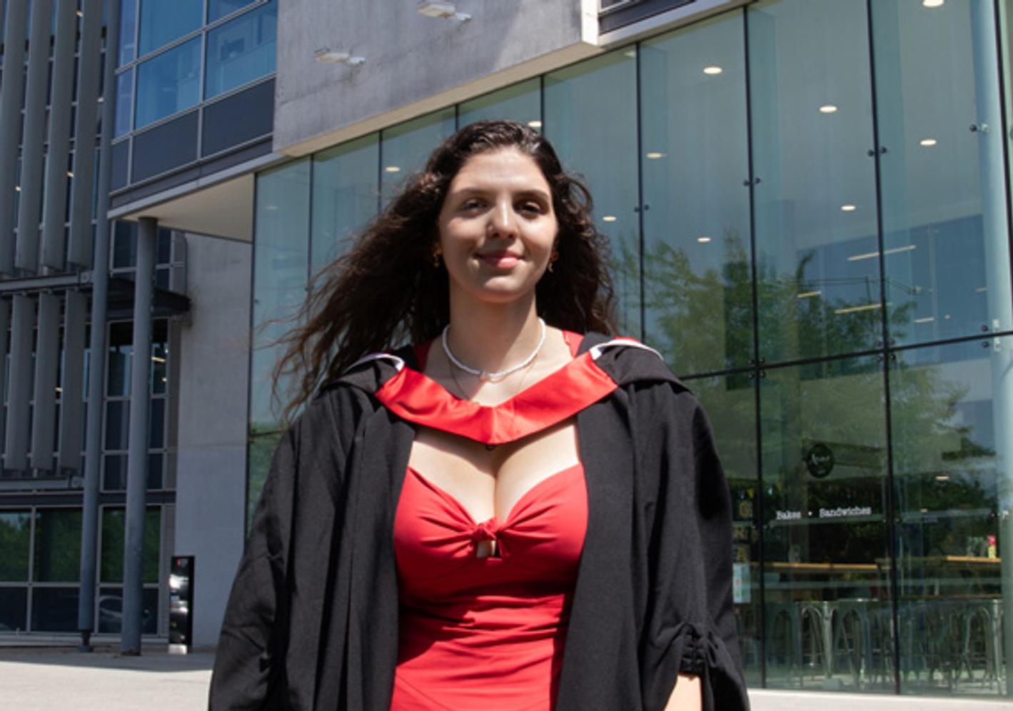 A female graduate in graduation gown posing in front of a glass building