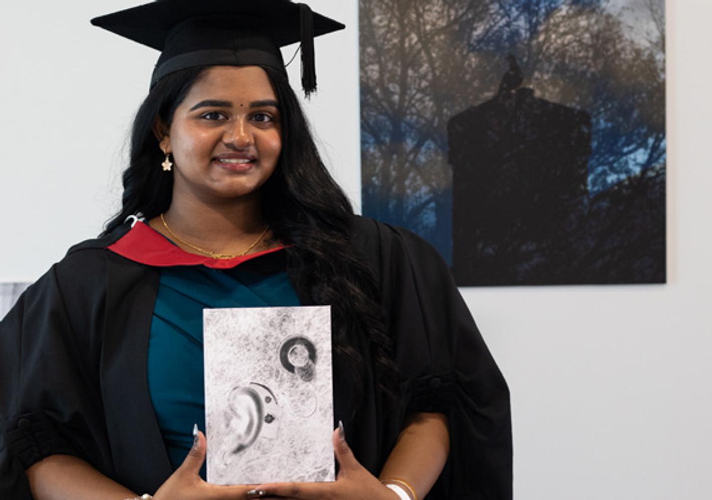 A woman in graduation gown poses with her certificate in front of a picture in a building