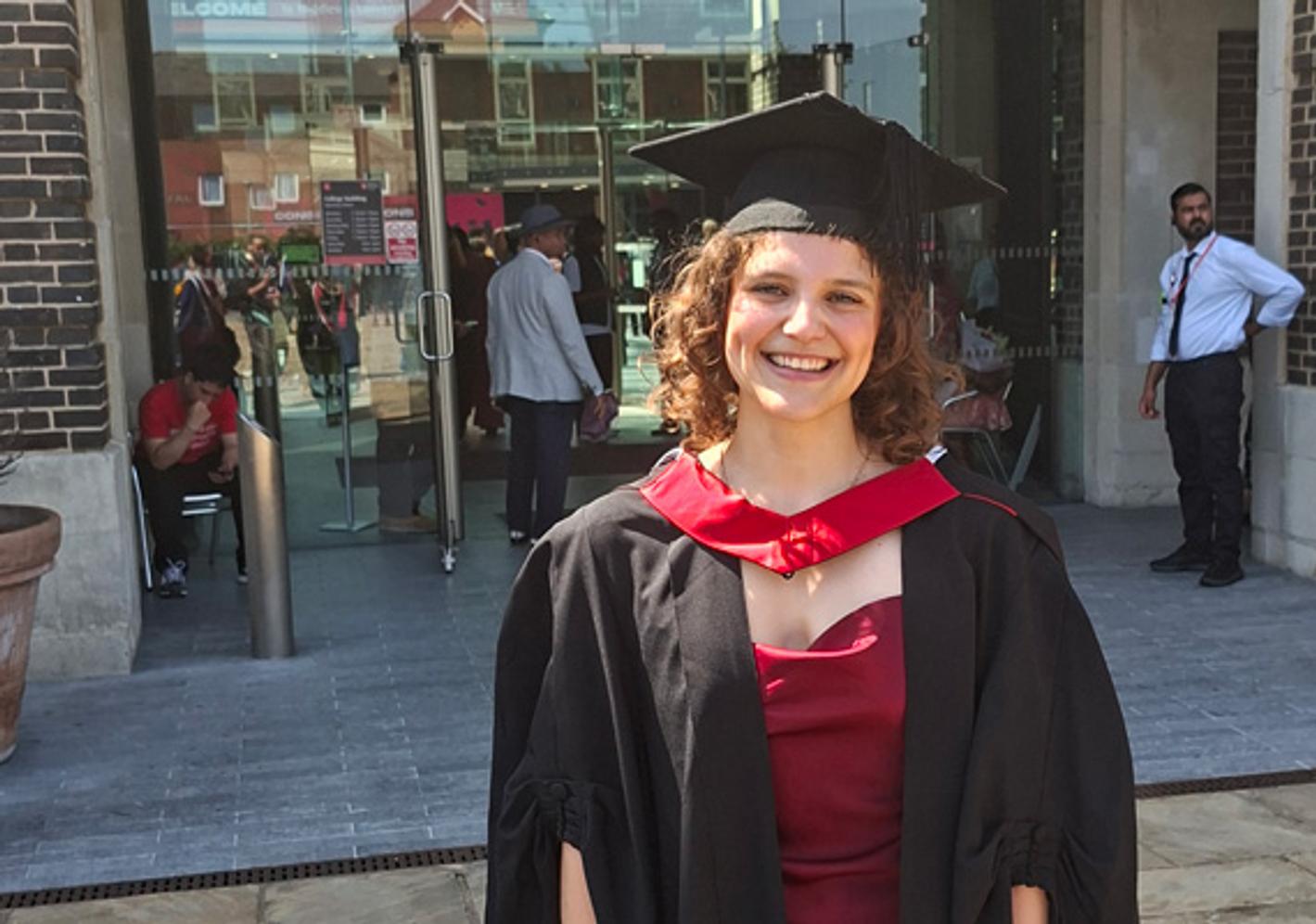 A woman in a graduation gown posing in front of a glass entrance to a university building
