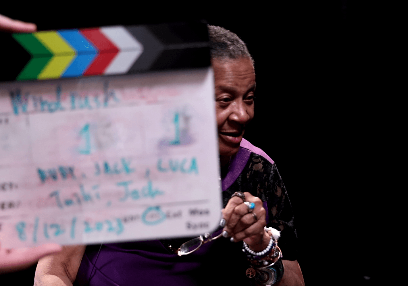 A woman smiling behind a film clapperboard that displays various production details. The board is titled 'Windrush' and includes names and a date, with the scene slightly out of focus.