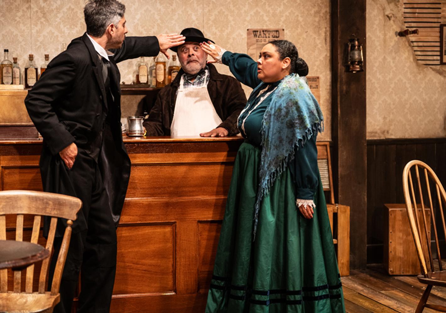 A man looks comically worried behind a bar as a man and a woman appear to touch his forehead to take his temperature