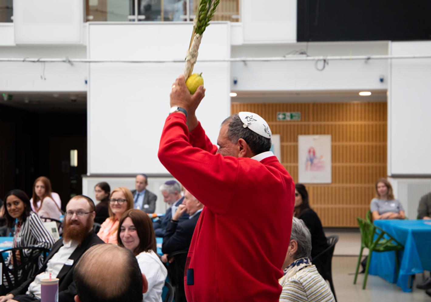 A jewish man wearing a red shirt raises food above his head to celebrate Sukkot
