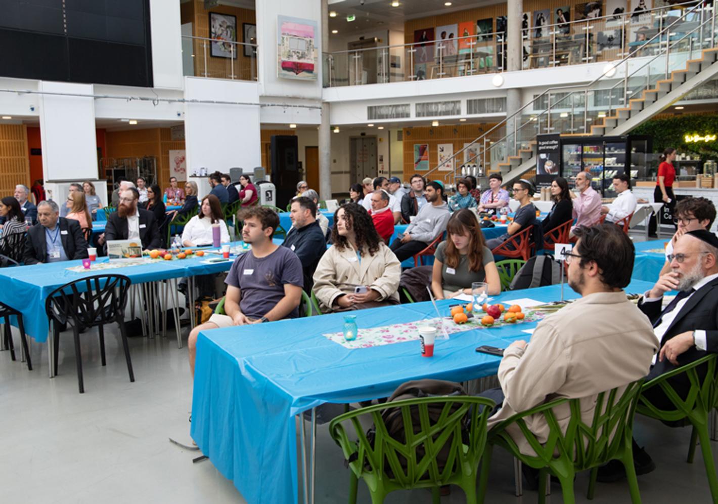 A large group of adults sitting at tables in a large hall listening to a speech
