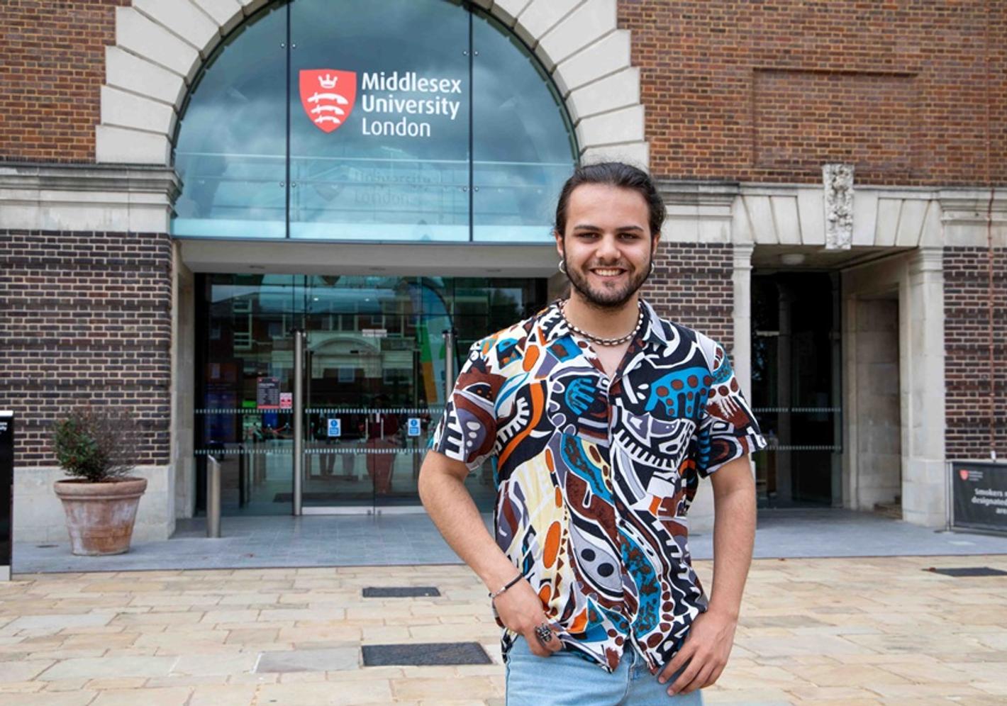 Omar Maaouan Veiga in a colourful shirt, standing and smiling outside the entrance to Middlesex University's College Building