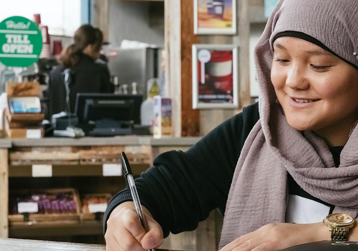 A sociologist woman wearing a headscarf sits at a table, engrossed in her work on a laptop.