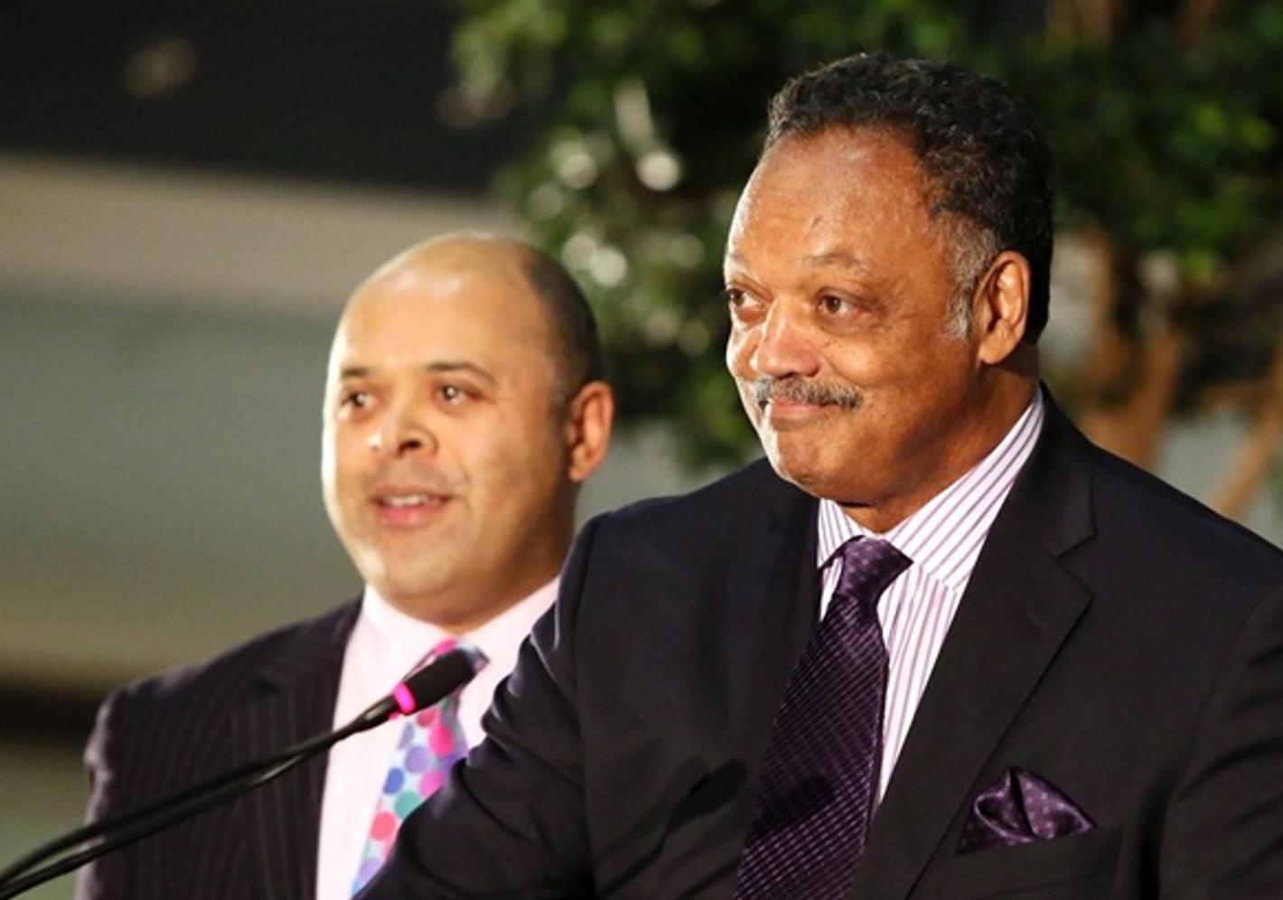 Jesse Jackson speaking at a podium, with Kurt Barling standing just behind him, both in smart suits