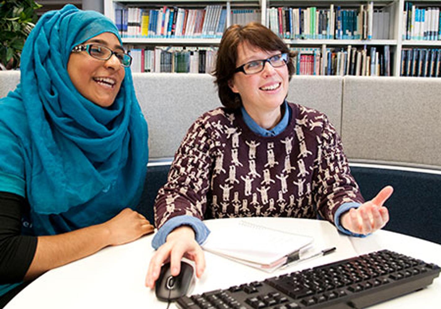 Two women sitting at a table, one typing on a computer while the other looks at the screen.