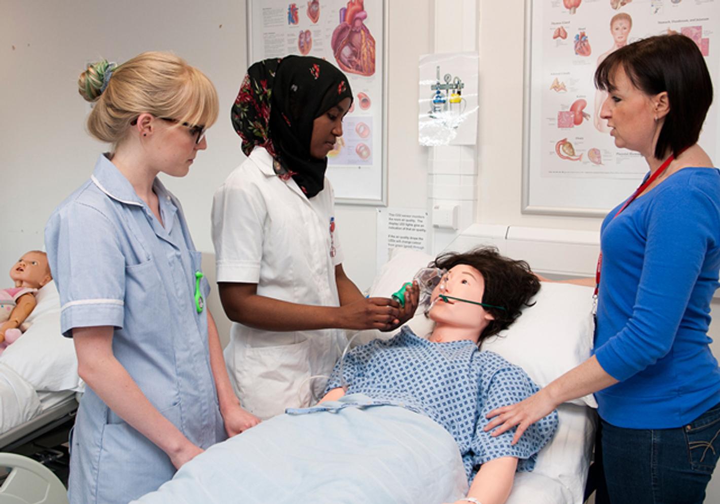 Two trainees nurses working with a dummy in a bed whilst being supervised by a tutor