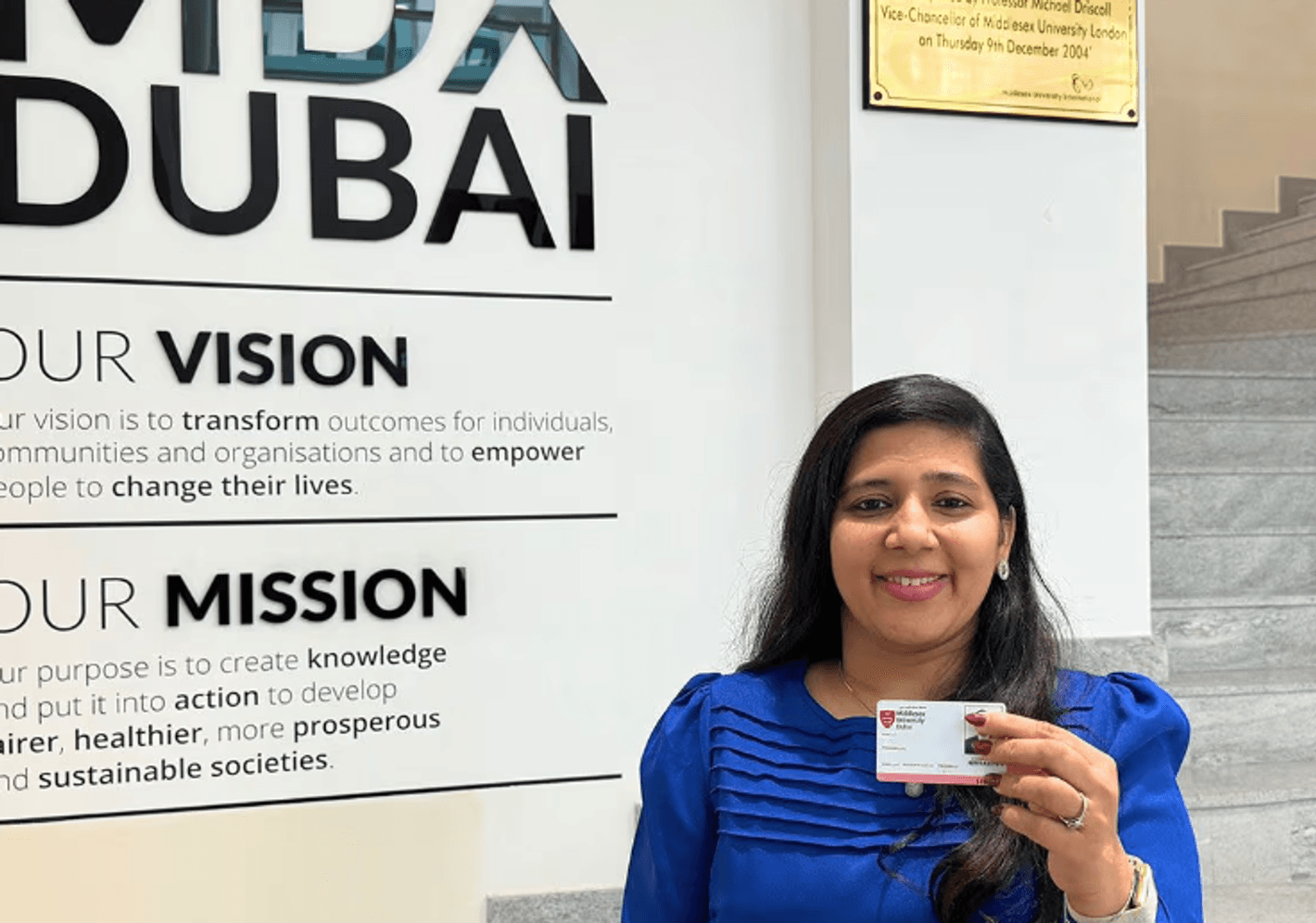 A smiling woman holding up her ID card in front of a sign for Middlesex University Dubai