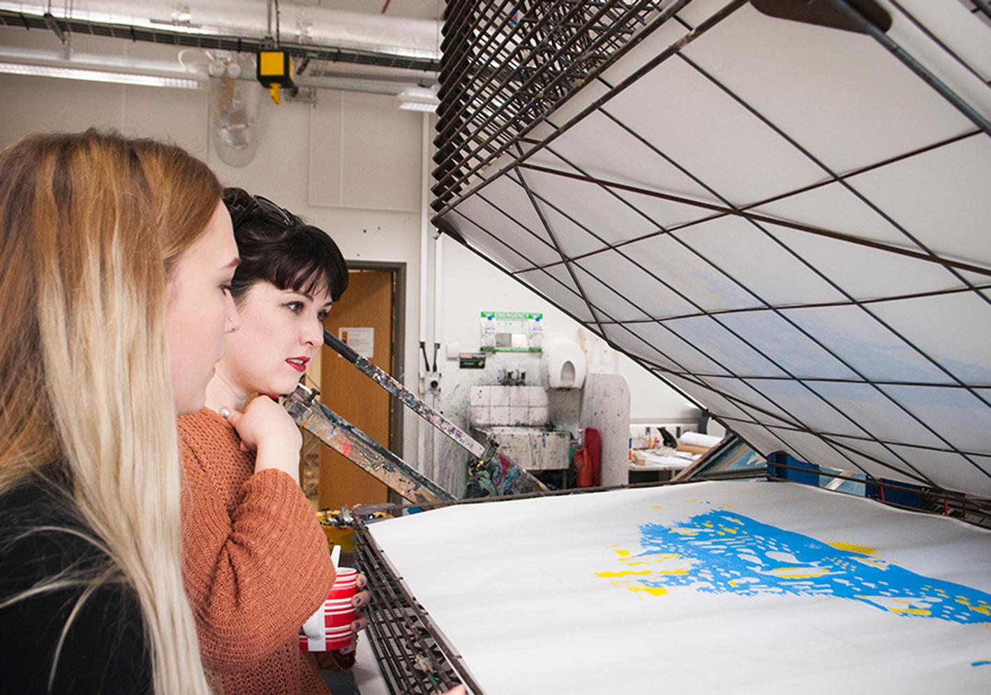 Two women at a Print Making Workshop examining a sizable sheet of paper, engrossed in their artistic endeavor.