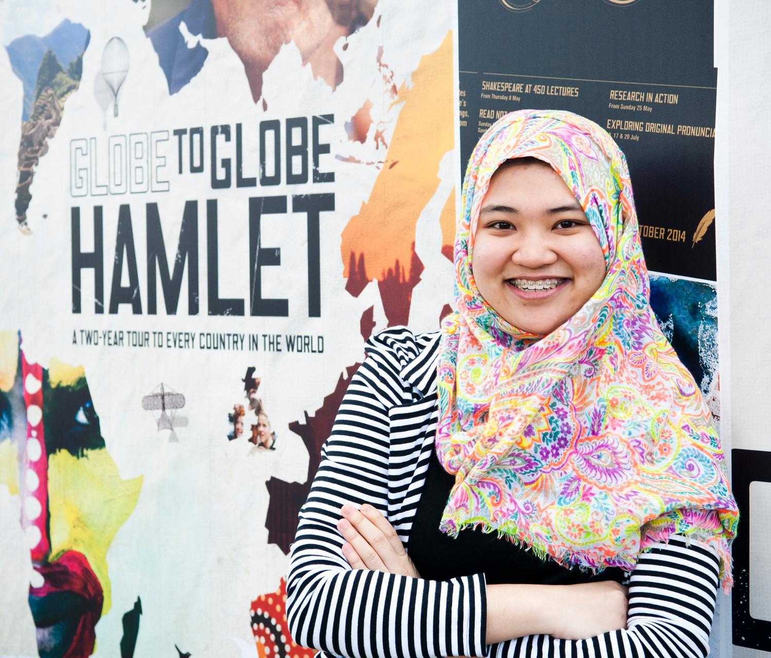 A woman wearing a headscarf stands in front of a poster promoting a visit to Shakespeare's Globe Theatre.