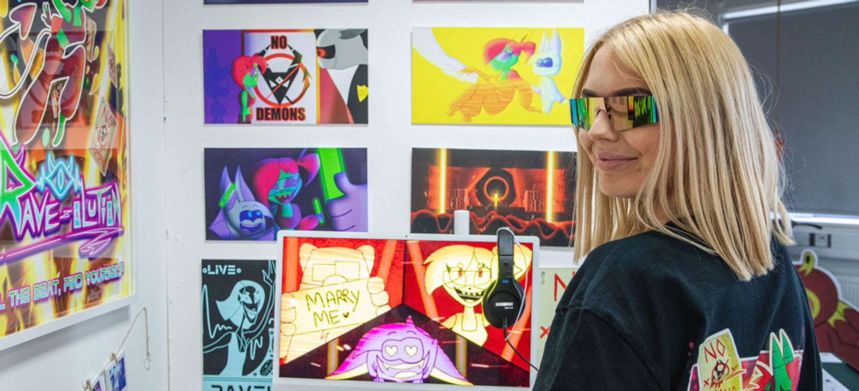 An female animation student with mirrored glasses amongst a colourful display of graphic work