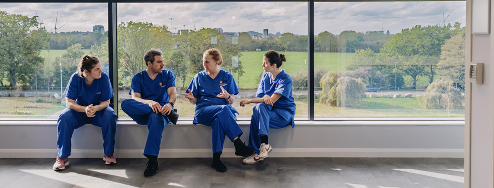 Medical staff in scrubs holding a discussion whilst sitting in a large window overlooking trees and a lawn