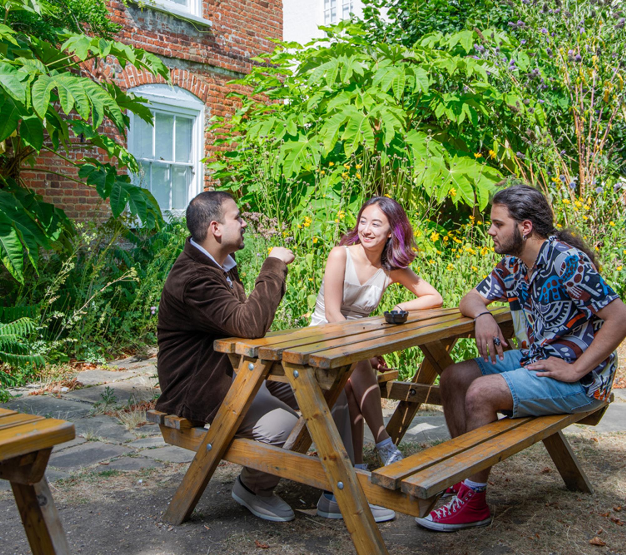 Three students sitting on a picnic bench surrounded by trees outside an old building
