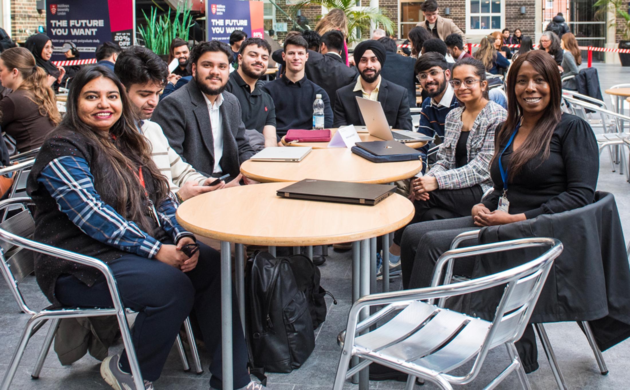 A large group of people sat round a table, smiling at the camera in a large university building
