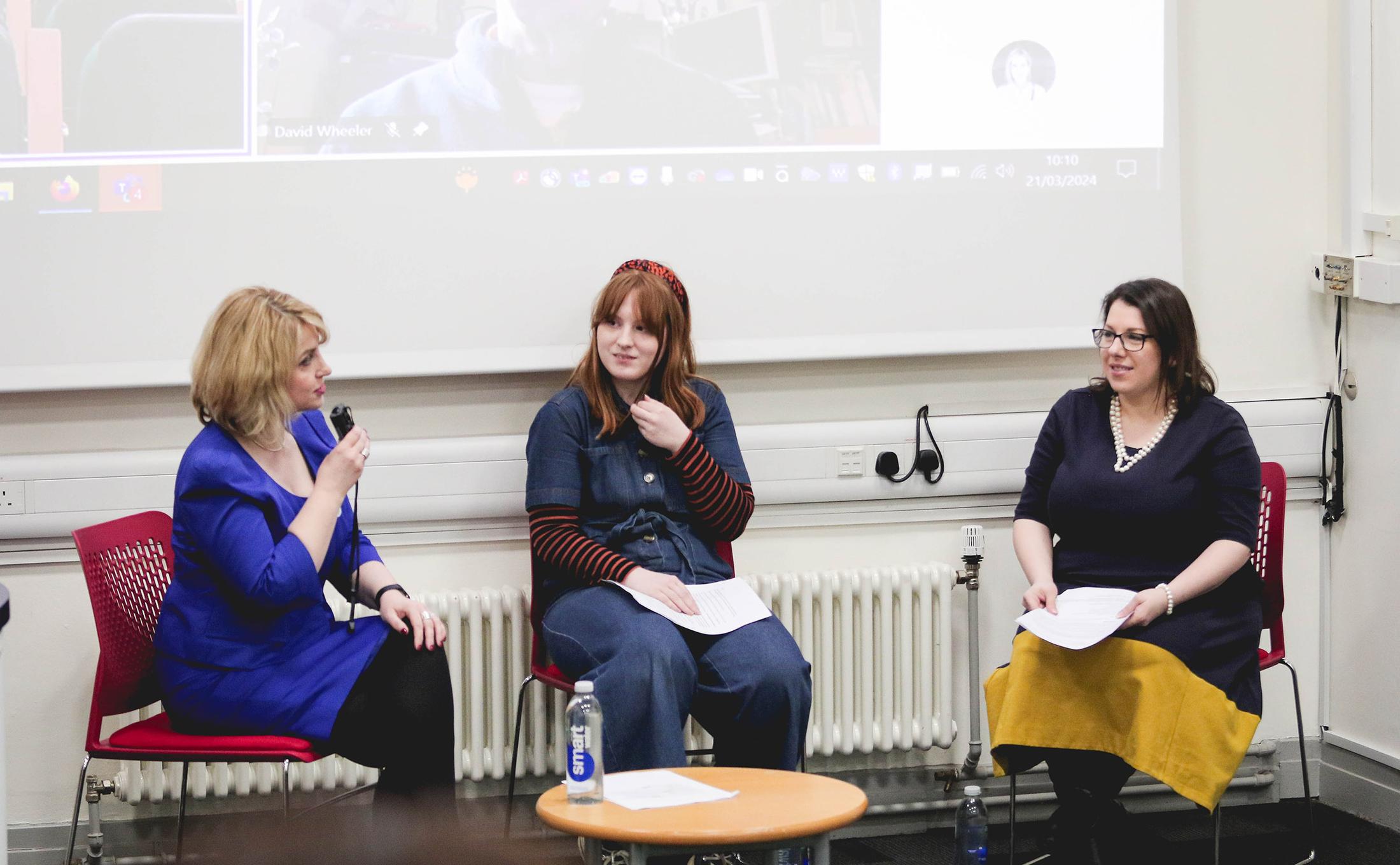 Three panellists having discussion with microphones, in front of screen with presentation