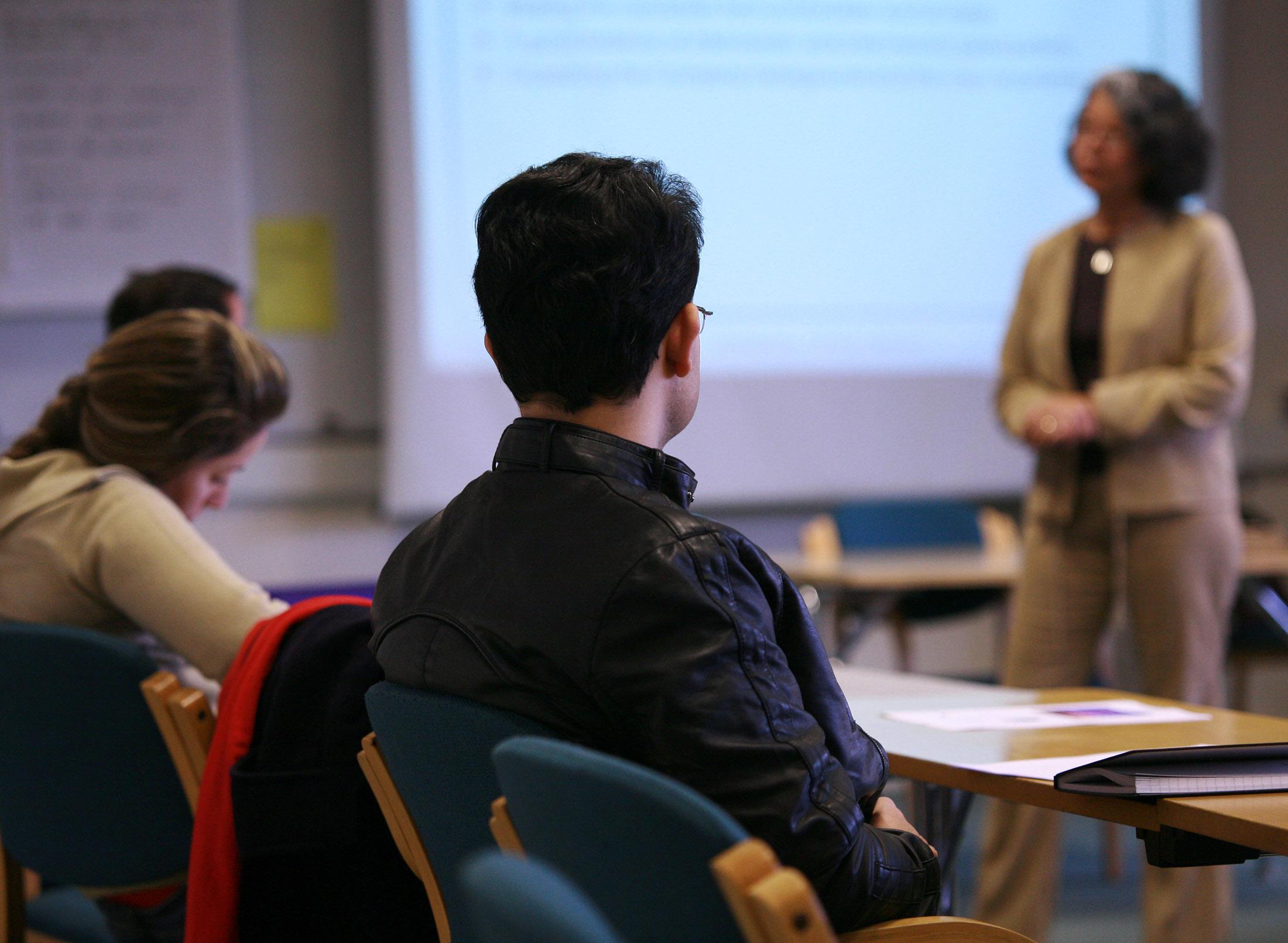 students listen to presentation, presenter stands in front of large display
