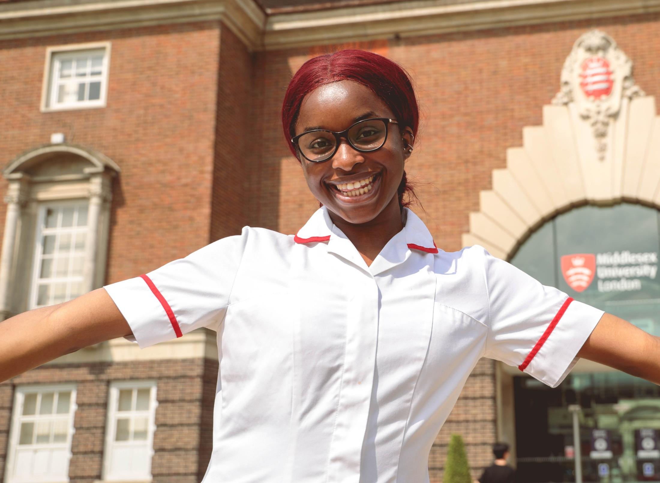 A smiling student in nurse's clothes holding her arms wide in front of the main entrance to Middlesex University London