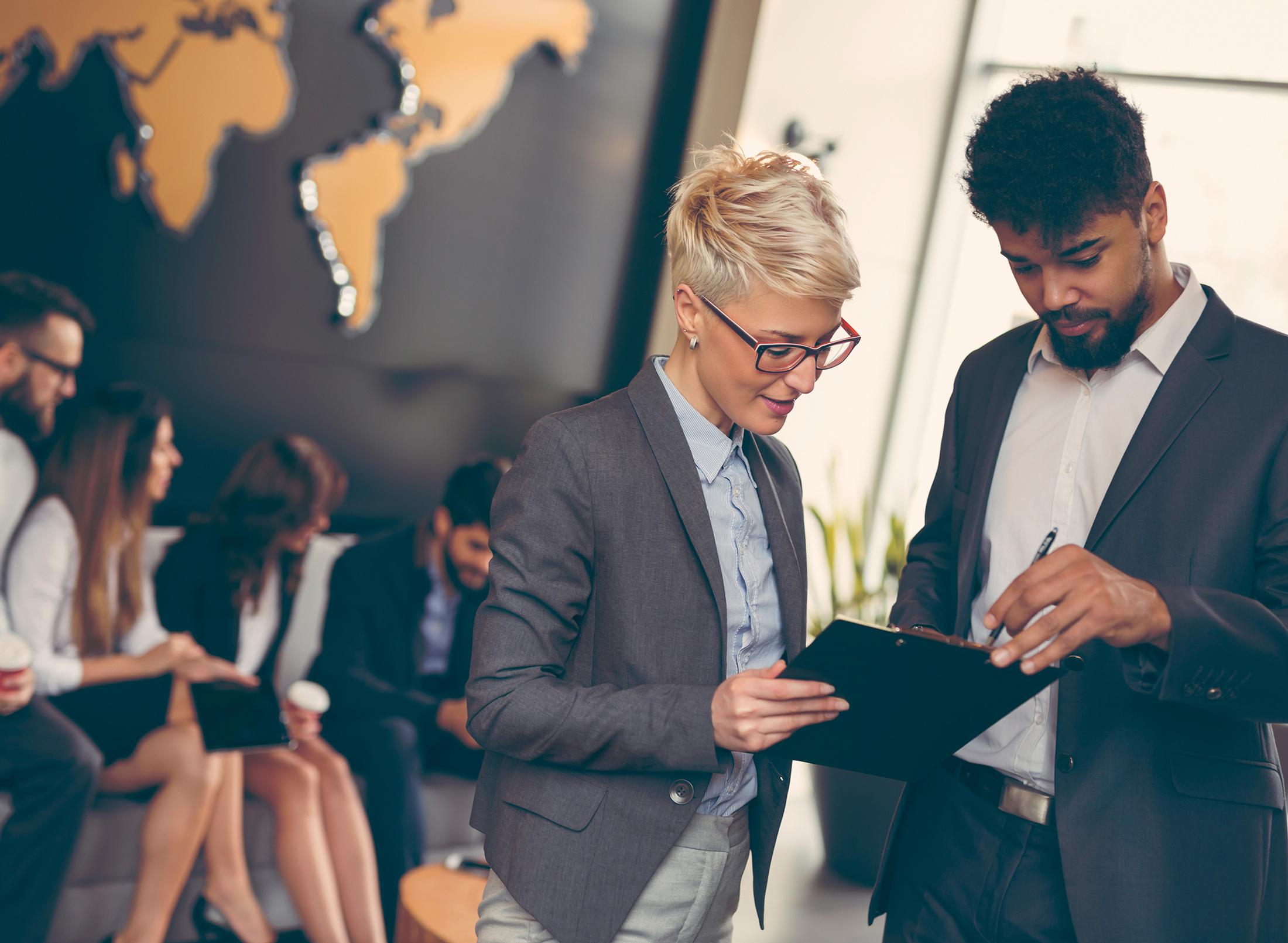 Two colleagues in an office look at a clipboard with colleague sat behind