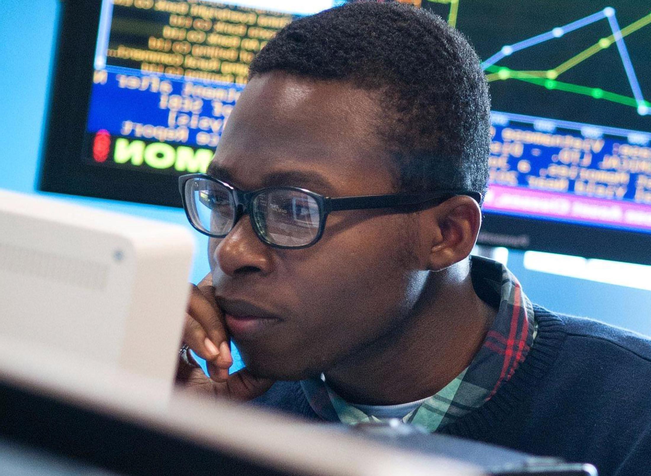 A student sitting looking at a computer screen, with a monitor displaying financial data behind him