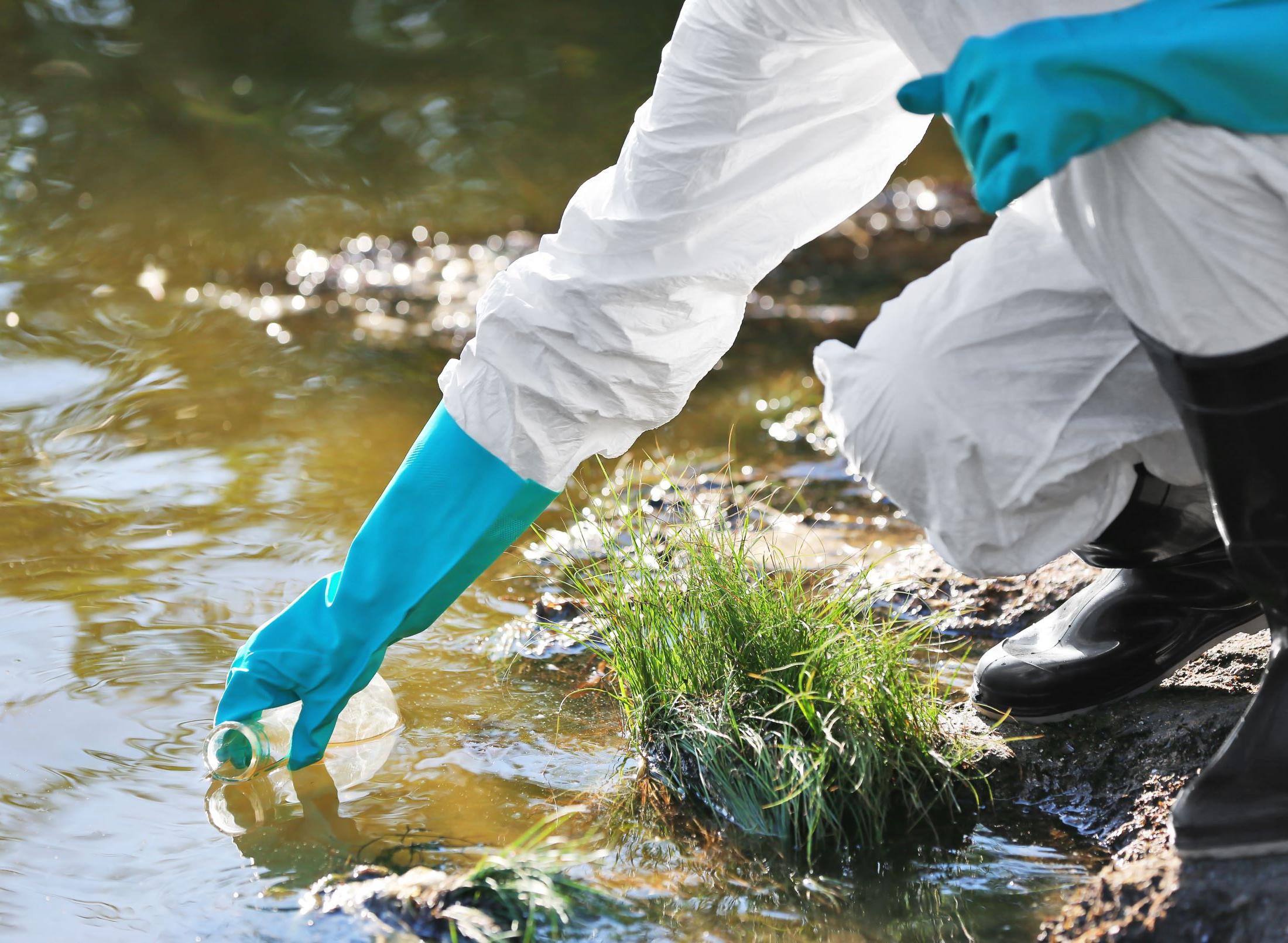 a person in white hazard suit with blue gloves reaches into the water to take a sample