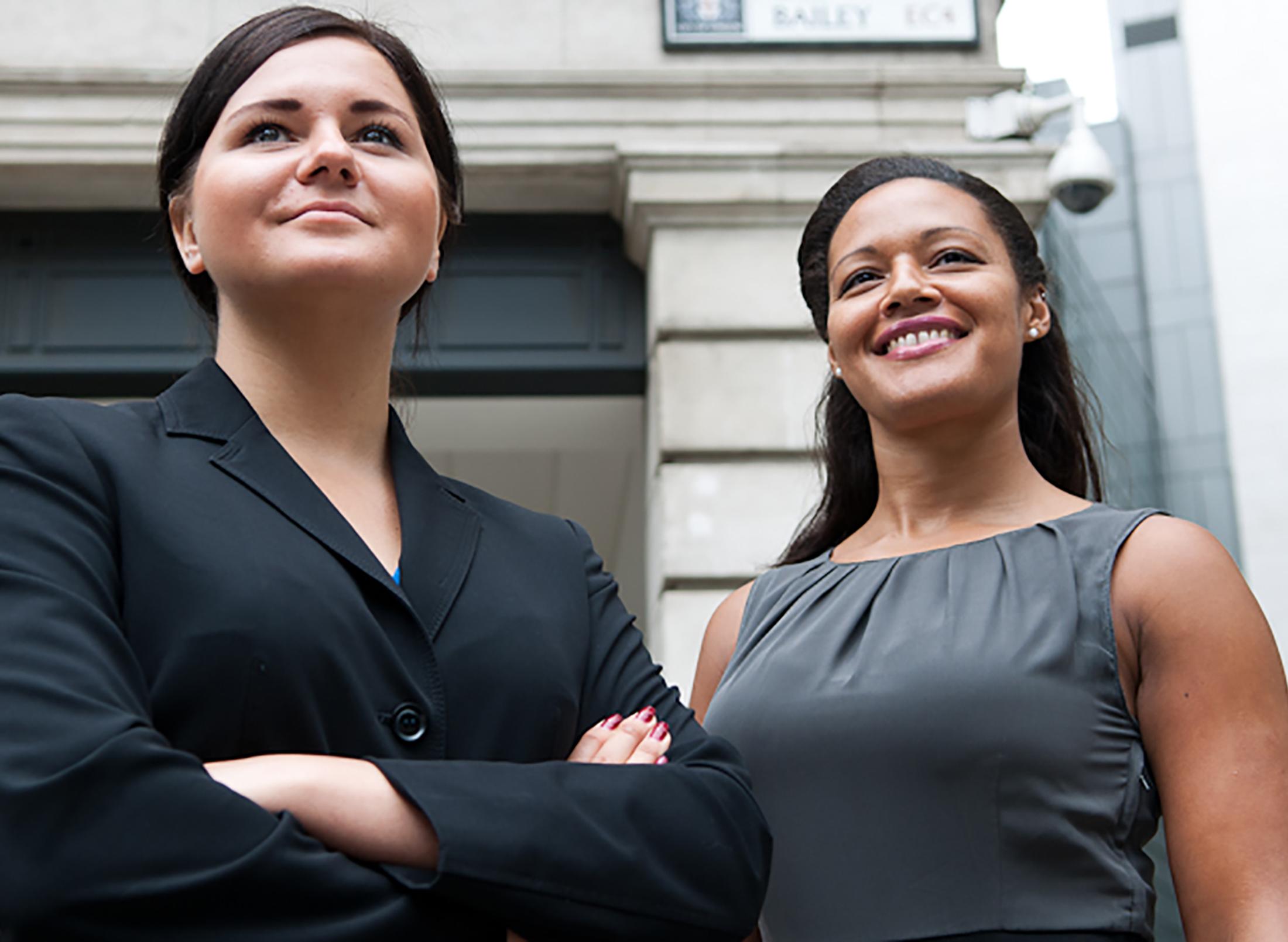 two smiling woman wearing business attire standing outside a building