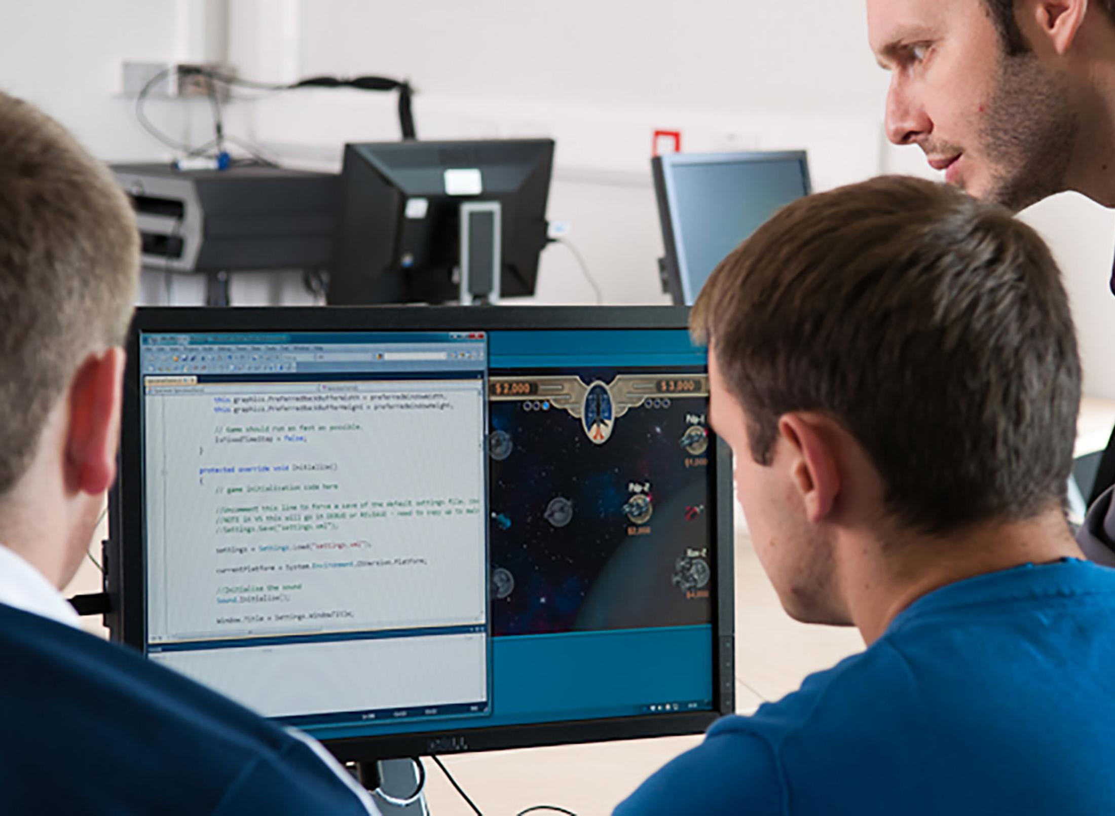 Two men sitting at a desk looking at a computer screen, with a third man standing next to them, leaning over to look at the screen