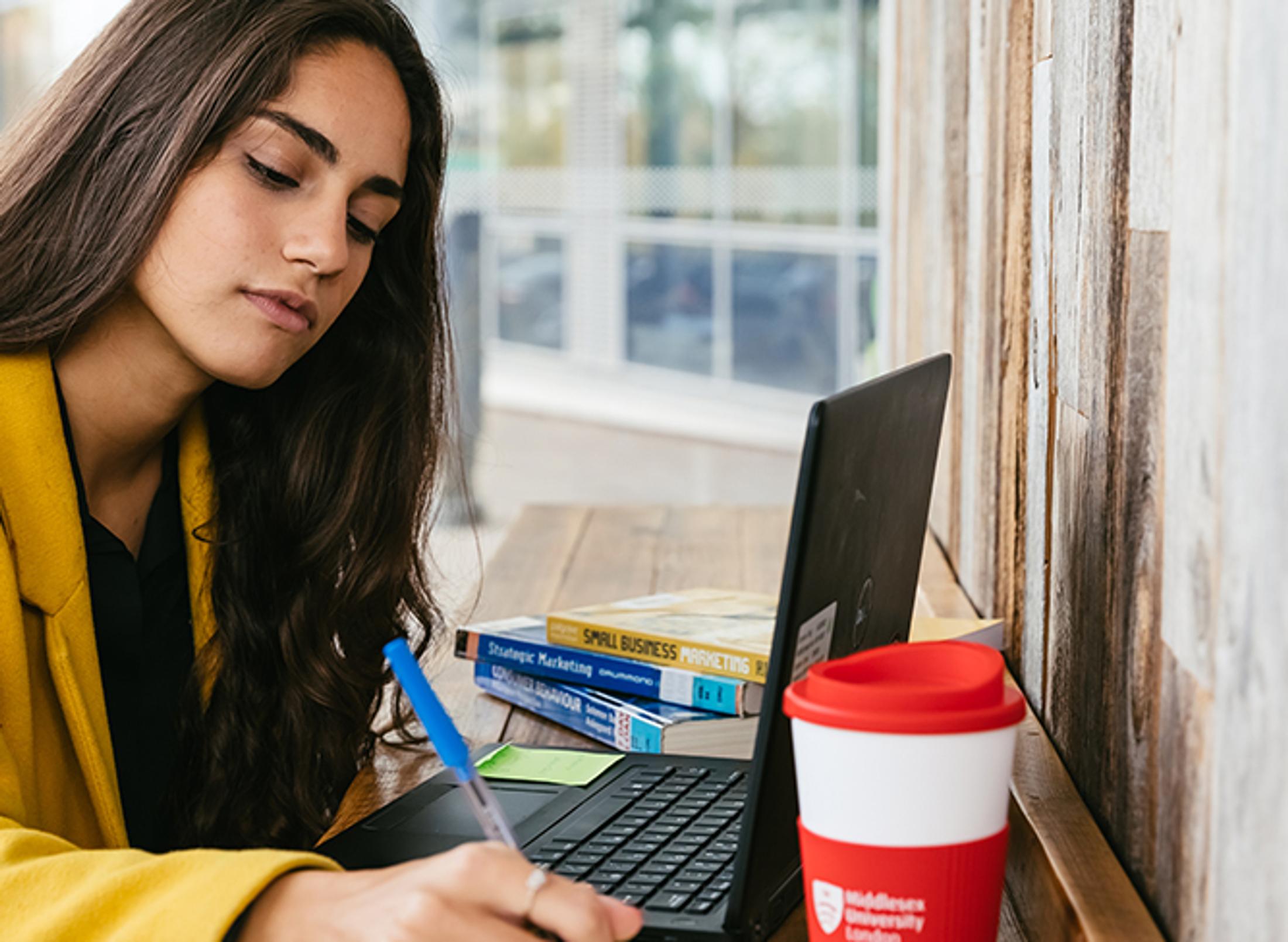 A woman sitting at a table, using a laptop and holding a cup of coffee.