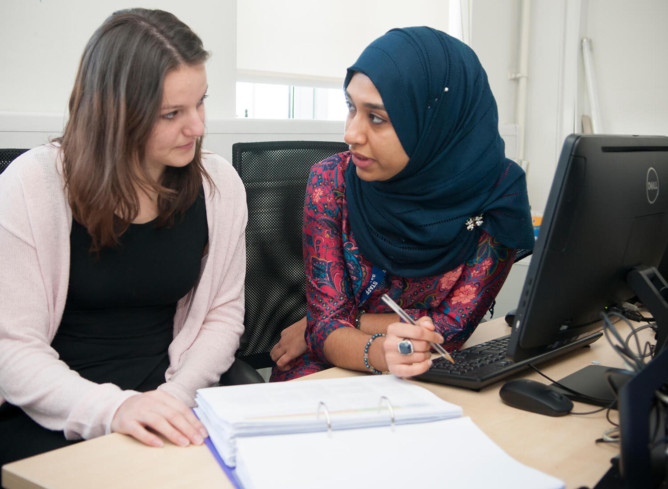 Two female students (one wearing a headscarf) sitting at a desk, talking