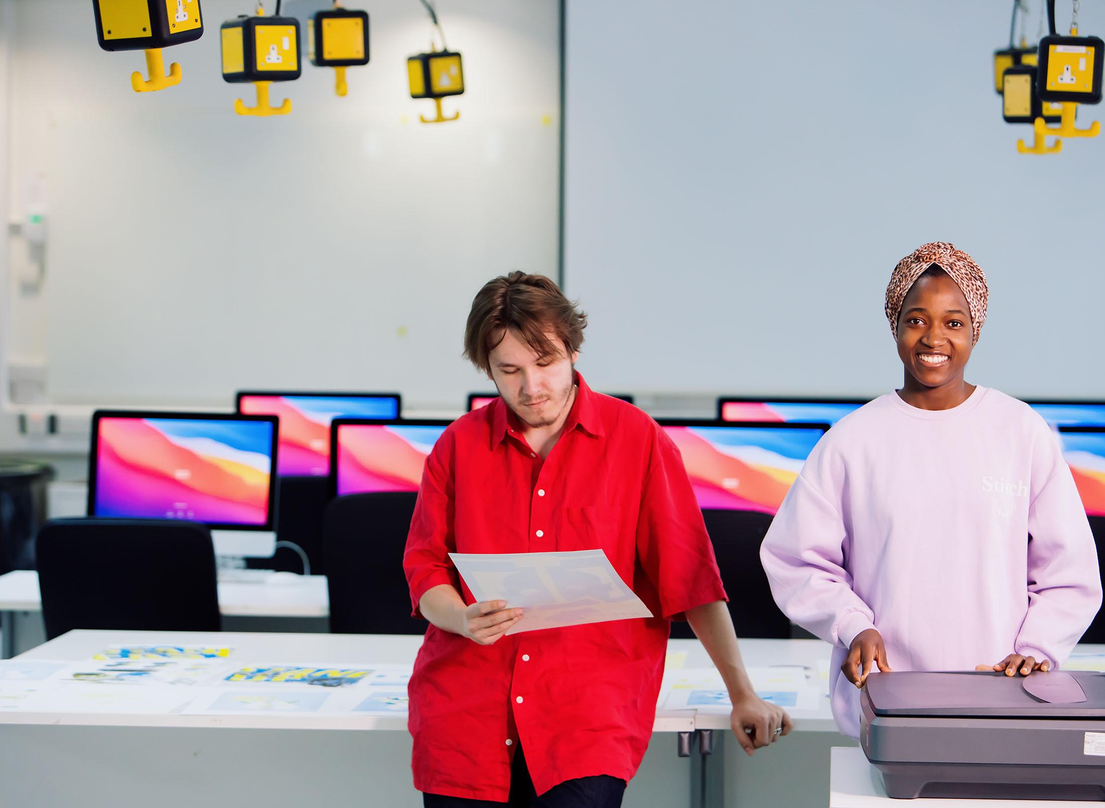 two students stand in studio full of design computers
