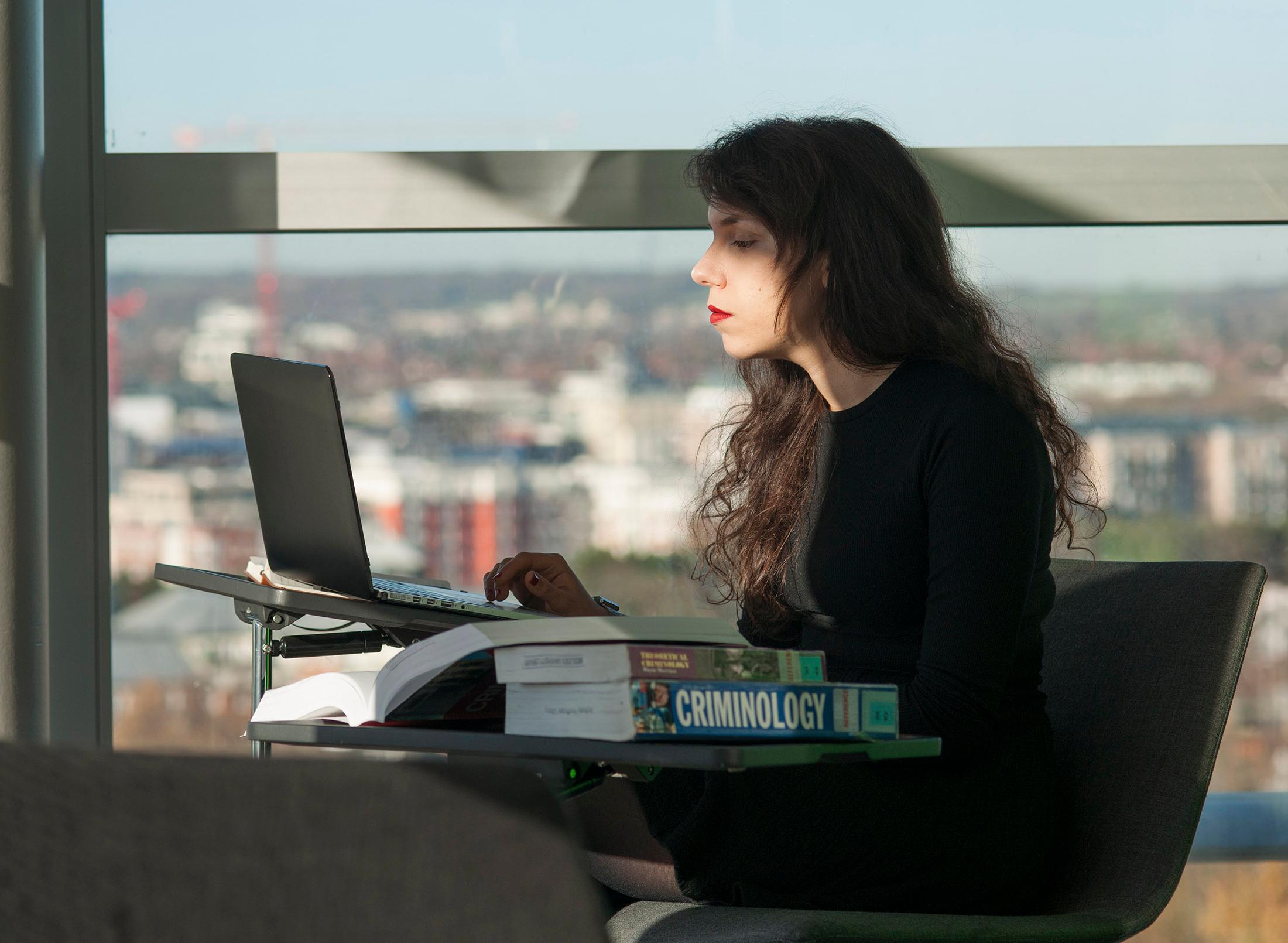 student concentrates at laptop in front of large window showing cityscape