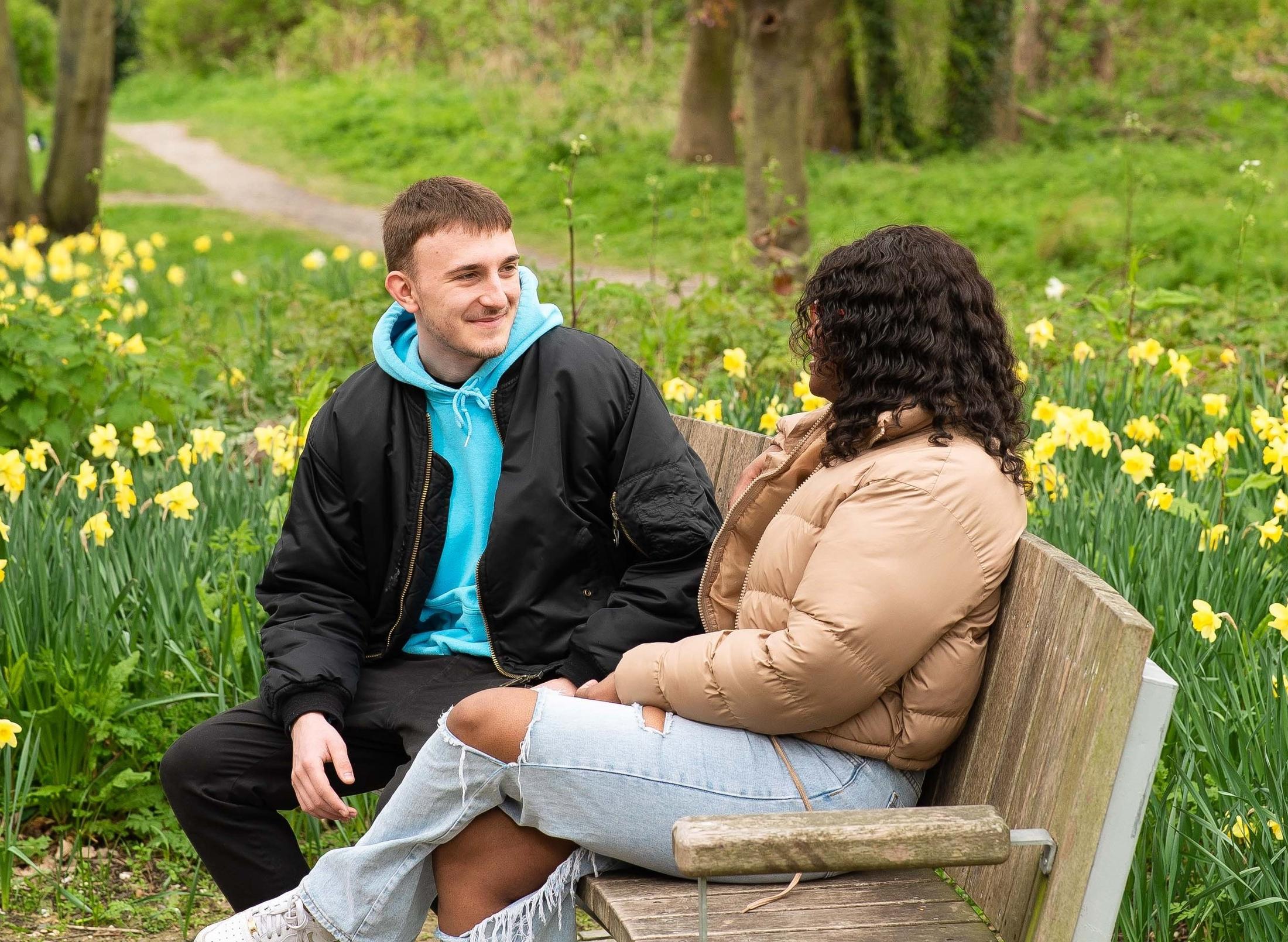 Two students sit on bench with green behind them and daffodils
