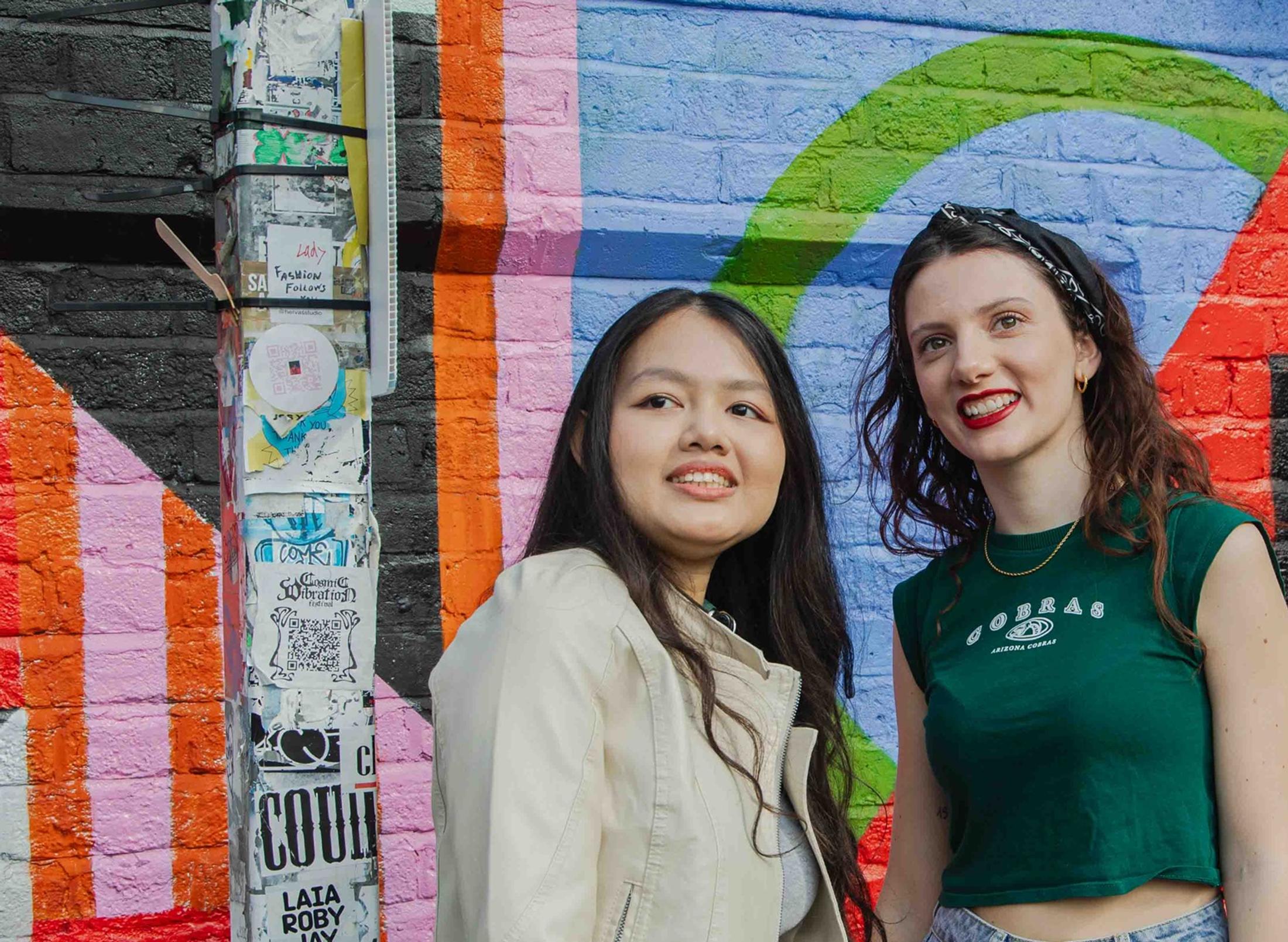 Two students smiling in front of a wall covered in artistic graffiti