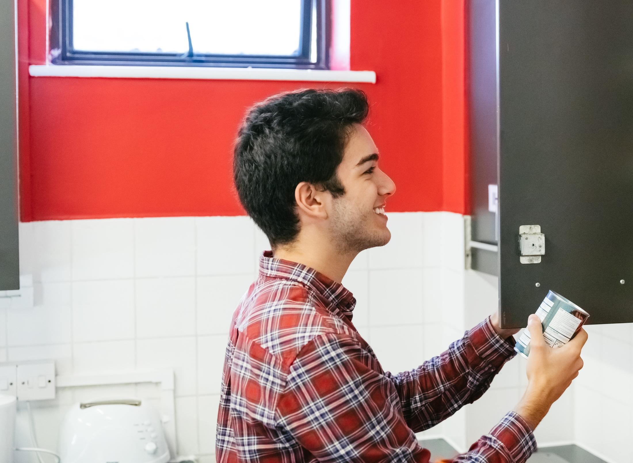 Student in kitchen holding a can of food with red wall in background.
