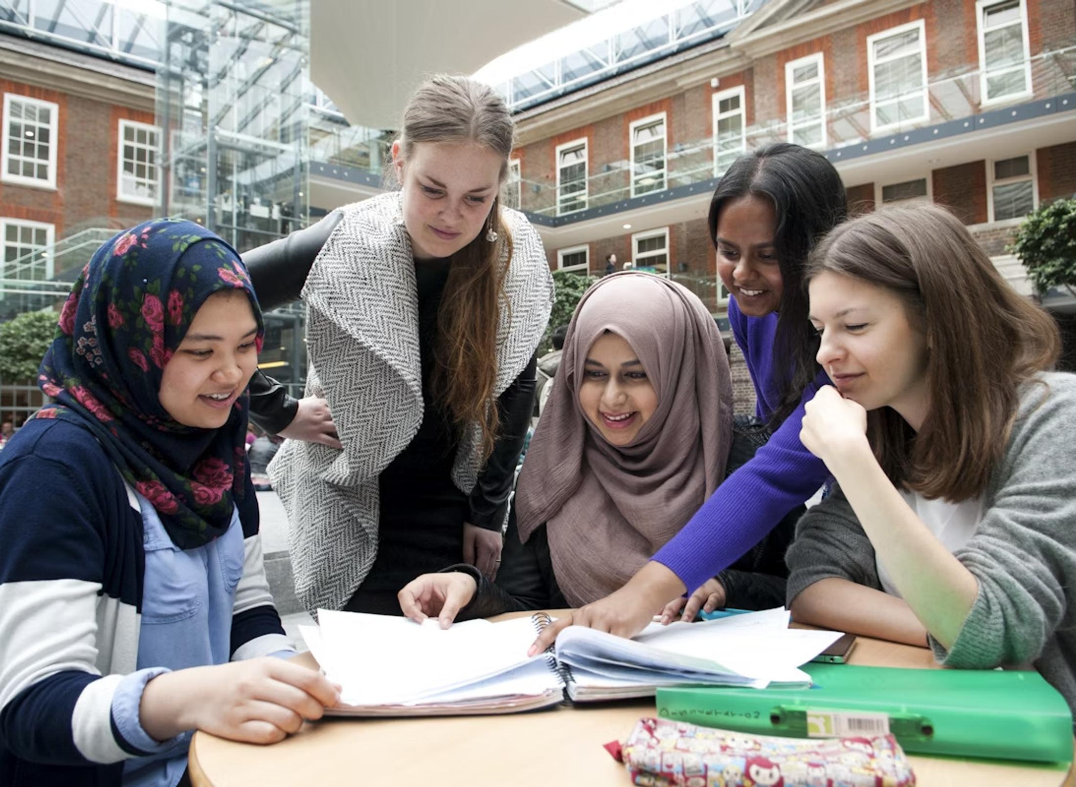 group of female middlesex university students gather around a table in the quad and look at notebooks together