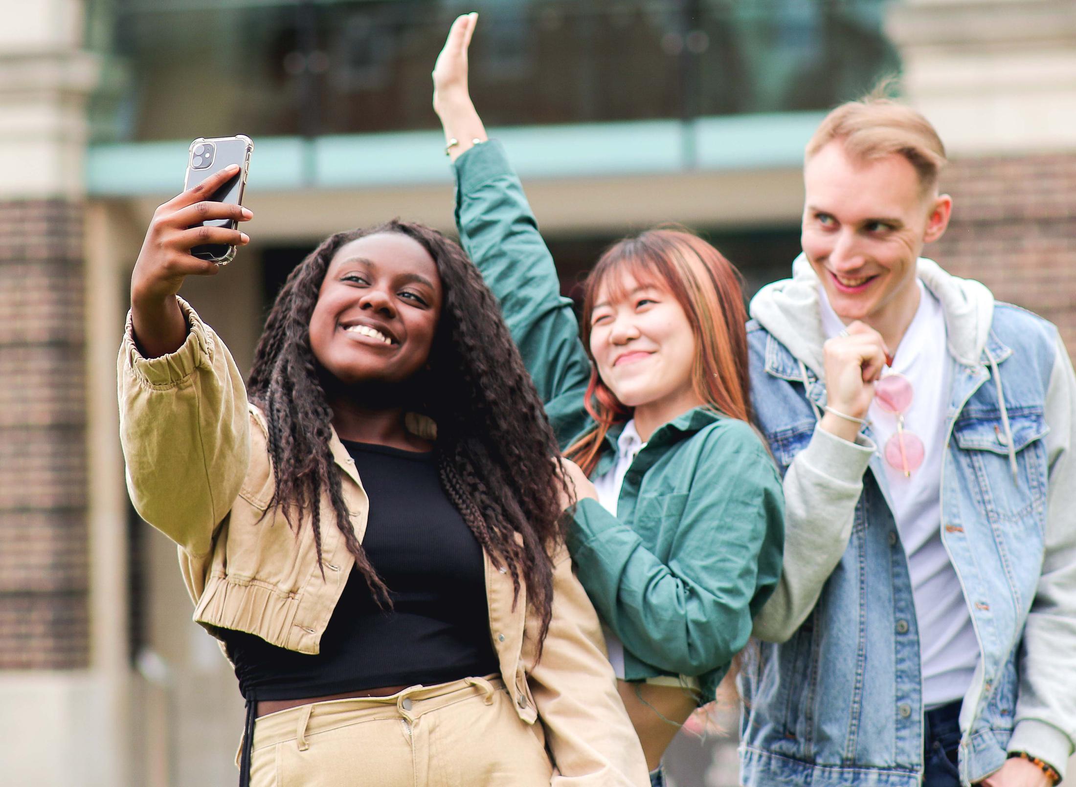 Three young people smiling and posing for a selfie in front of a university building.