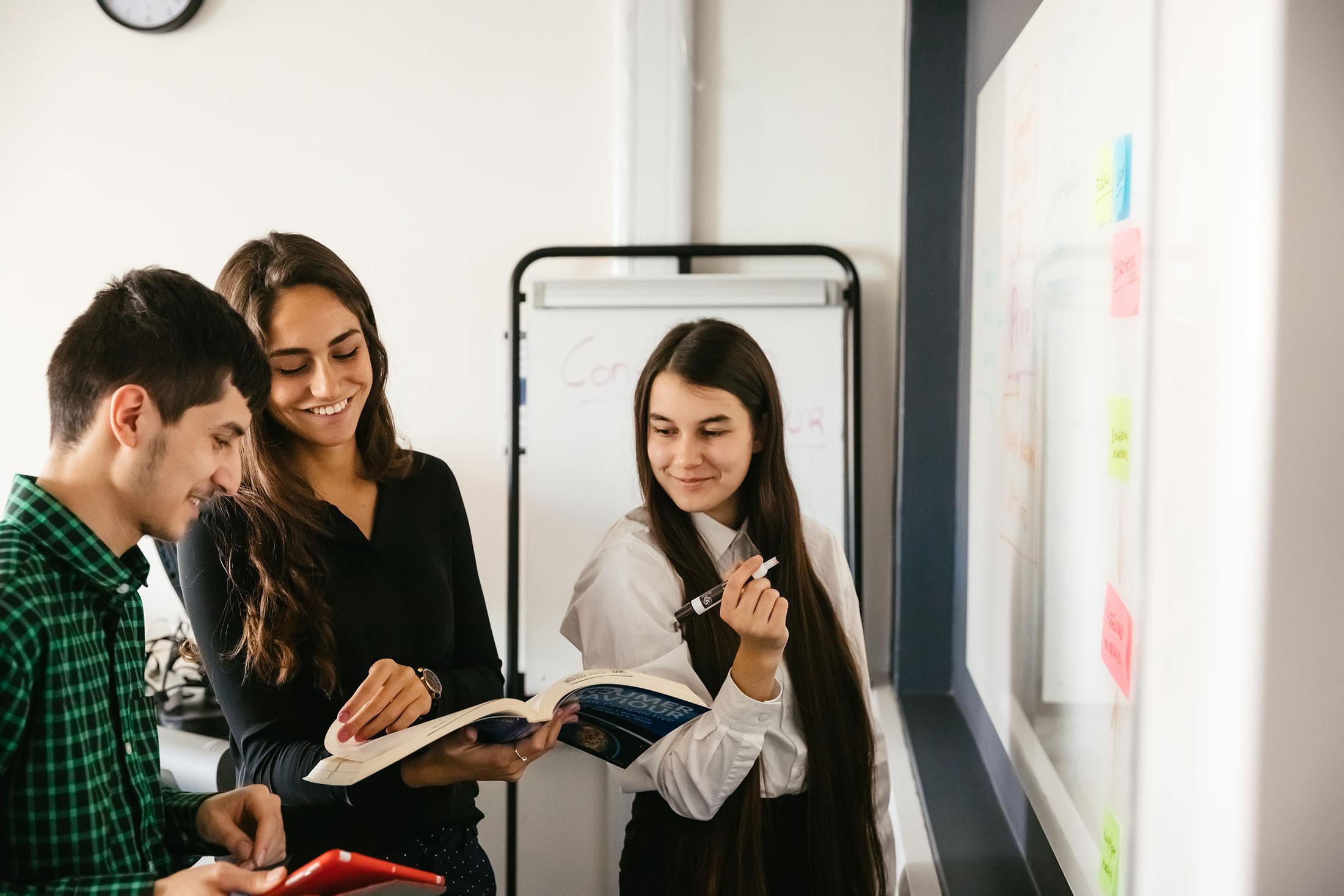 two students look at notebooks, while another prepares to write on whiteboard