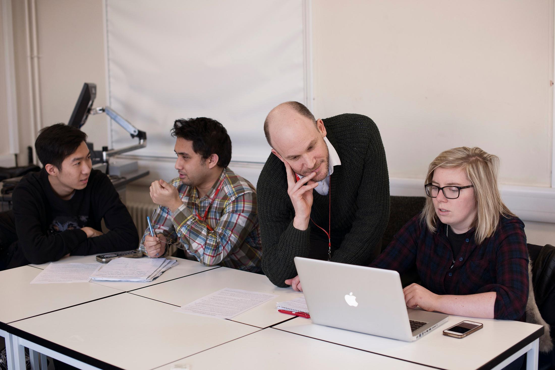 Group of people working informally at laptops, one stands to look at anothers computer