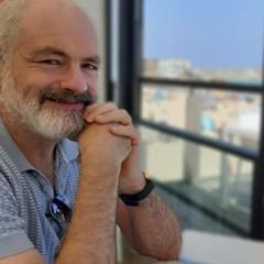 A man with a beard sat at a table, looking towards the camera with a window view in the background