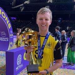 A smiling young man in sports kit holds a trophy bedecked in champions