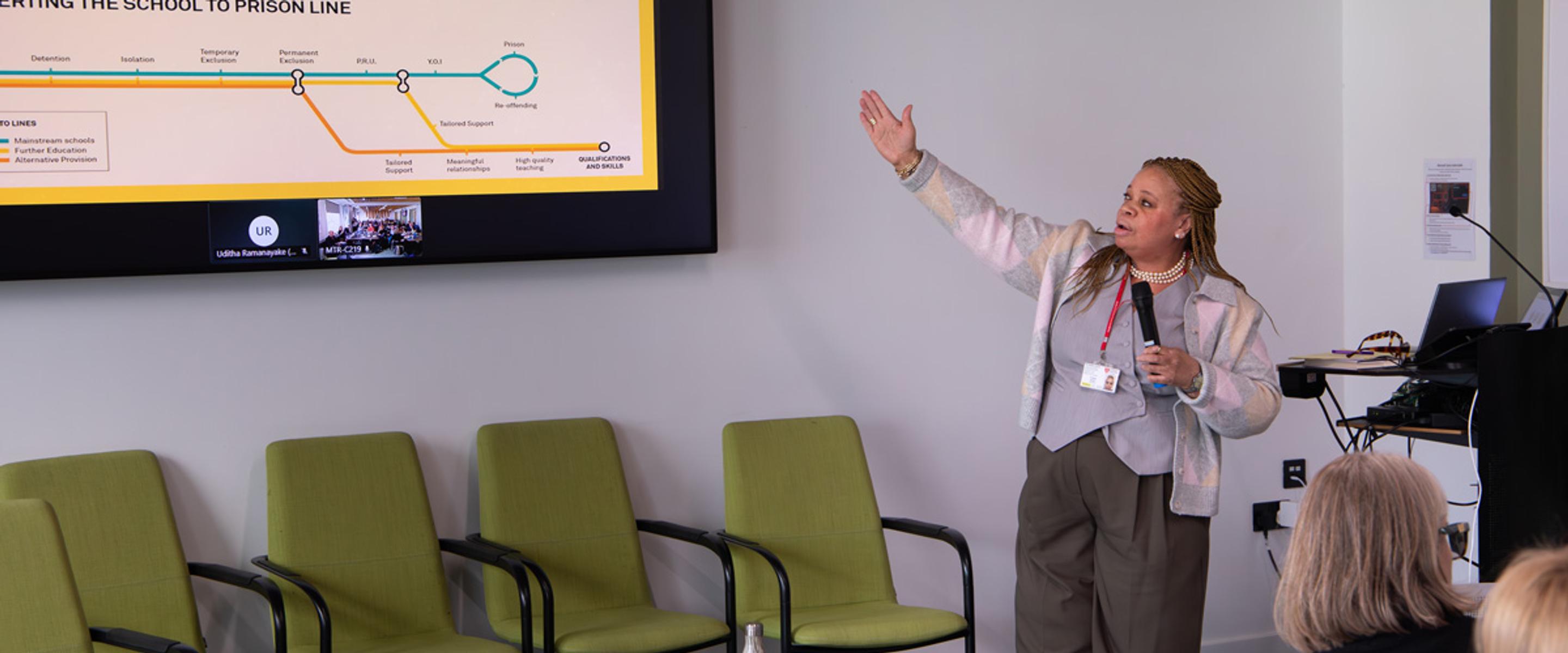 A woman delivering a lecture referring to a slide on a large screen whilst holding a microphone