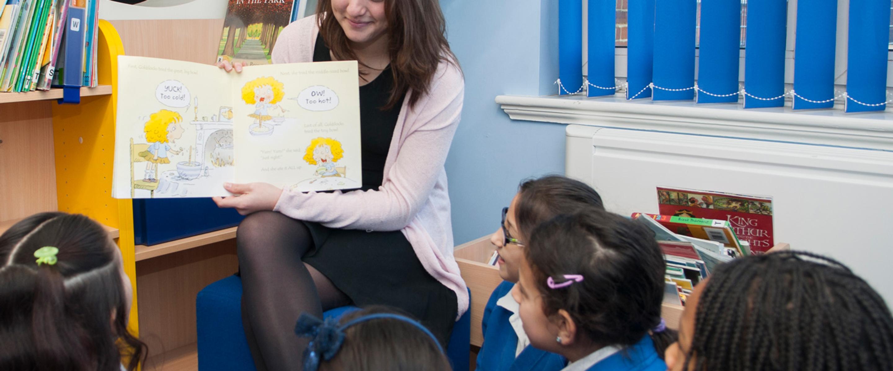 A teacher reads a picture book to a group of primary school children seated in a library.
