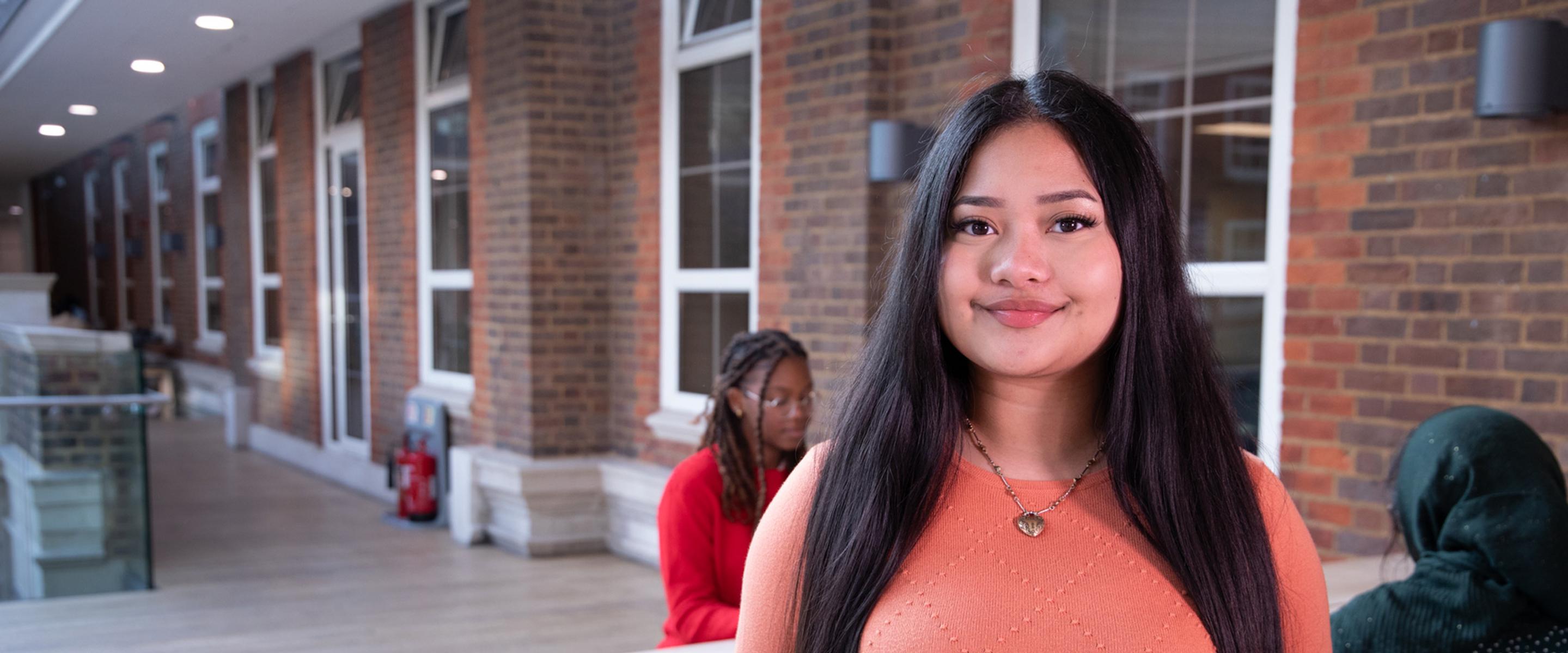 Female student stood on campus with students sat behind